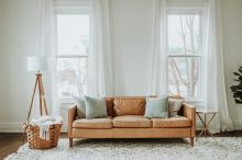 Sofa vor zwei großen Fenstern, auf hellem Teppich, links eine Stehlampe und ein Korb mit Decke.